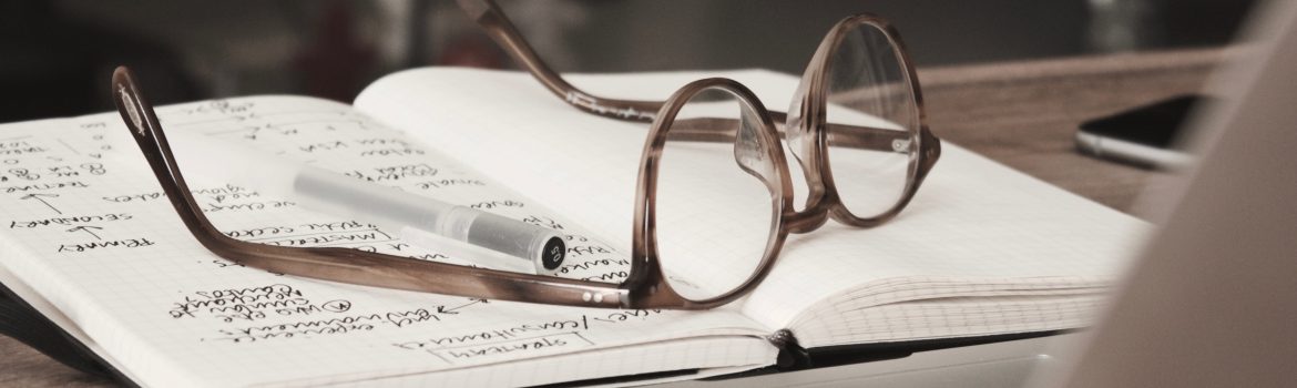 Glasses lying on the notes with pen on it. A wooden table and an Iphone.