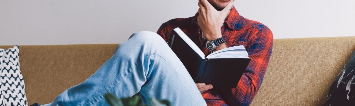 A man smiling and reading a book of a famous author sitting on a sofa.