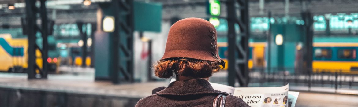 Woman in a hat reading a newspaper on a train station.