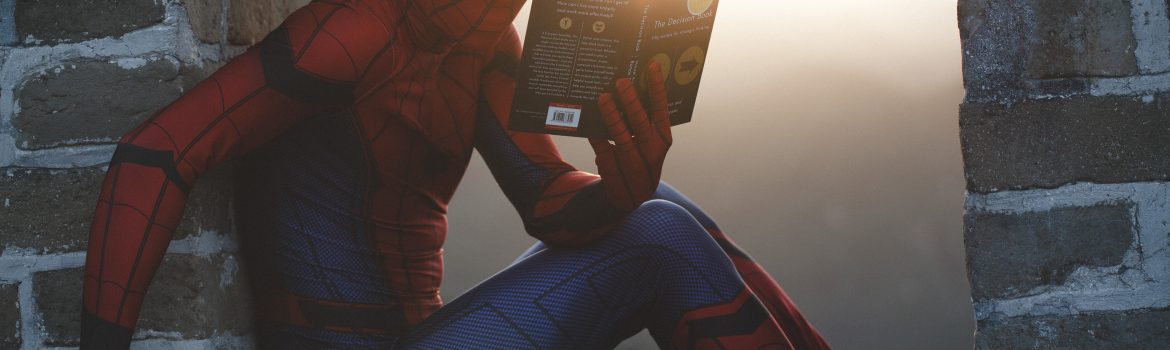 Spiderman is reading a book about "Decisions" sitting on a wall of rocks.