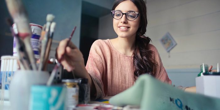 A girl in a pink sweater, wearing glasses is painting in a room.