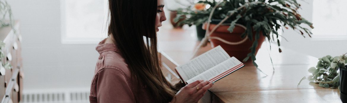 A girl in a pink shirt reading a book in the library with plants on bookcases.