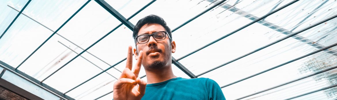 Man in a blue t-shirt and glasses making a peace sign with his hand.